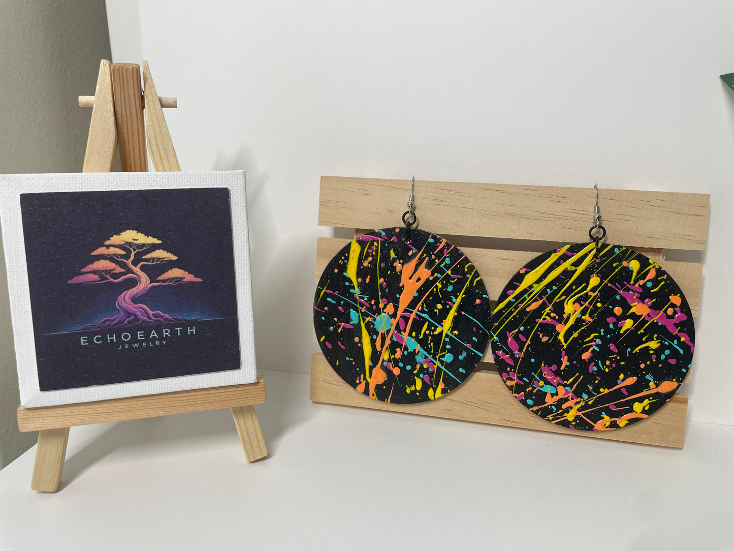 Colorful splatter-patterned earrings on a wooden stand with a small framed artwork on a white surface.