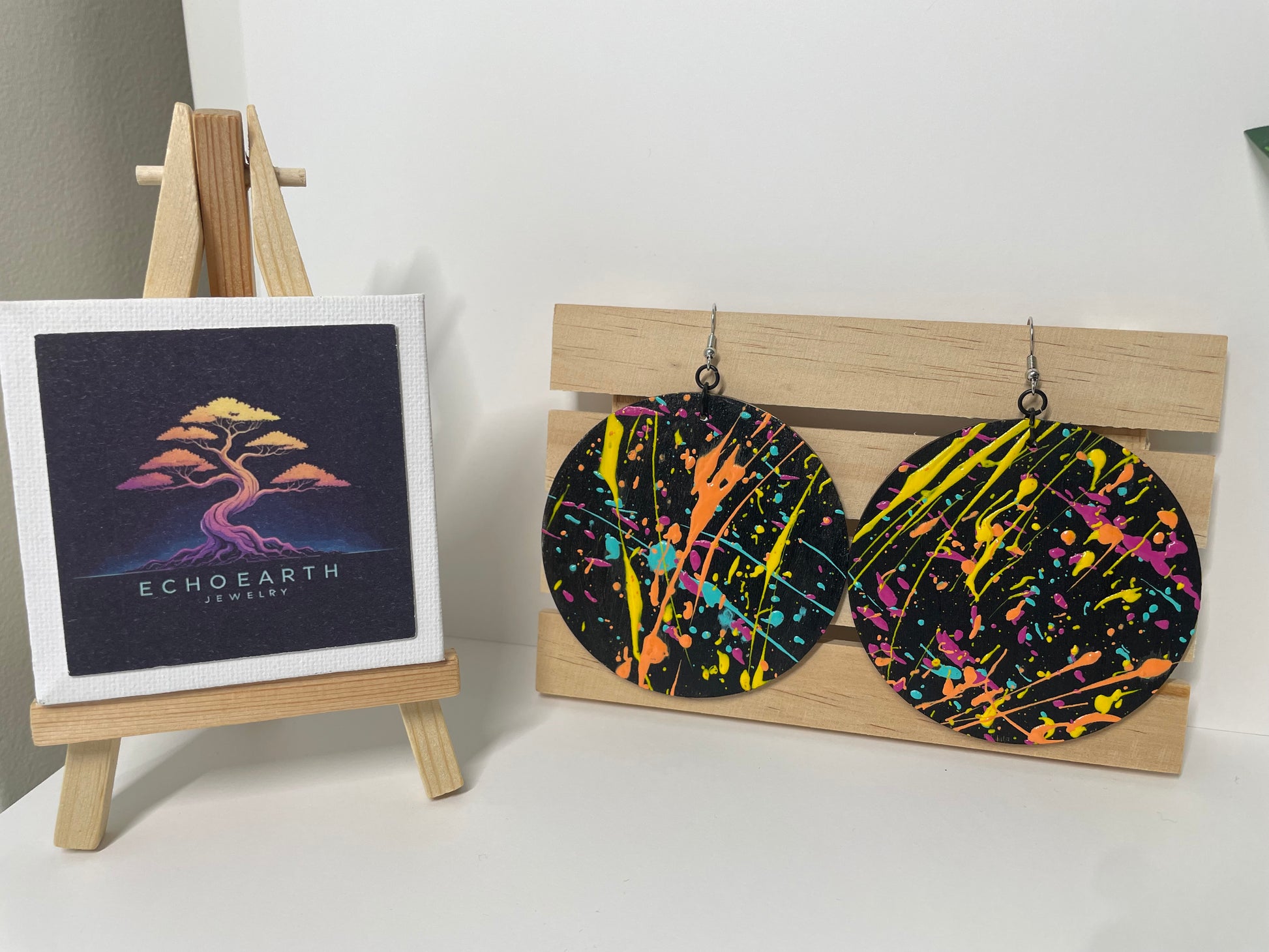 Colorful splatter-patterned earrings on a wooden stand with a small framed artwork on a white surface.