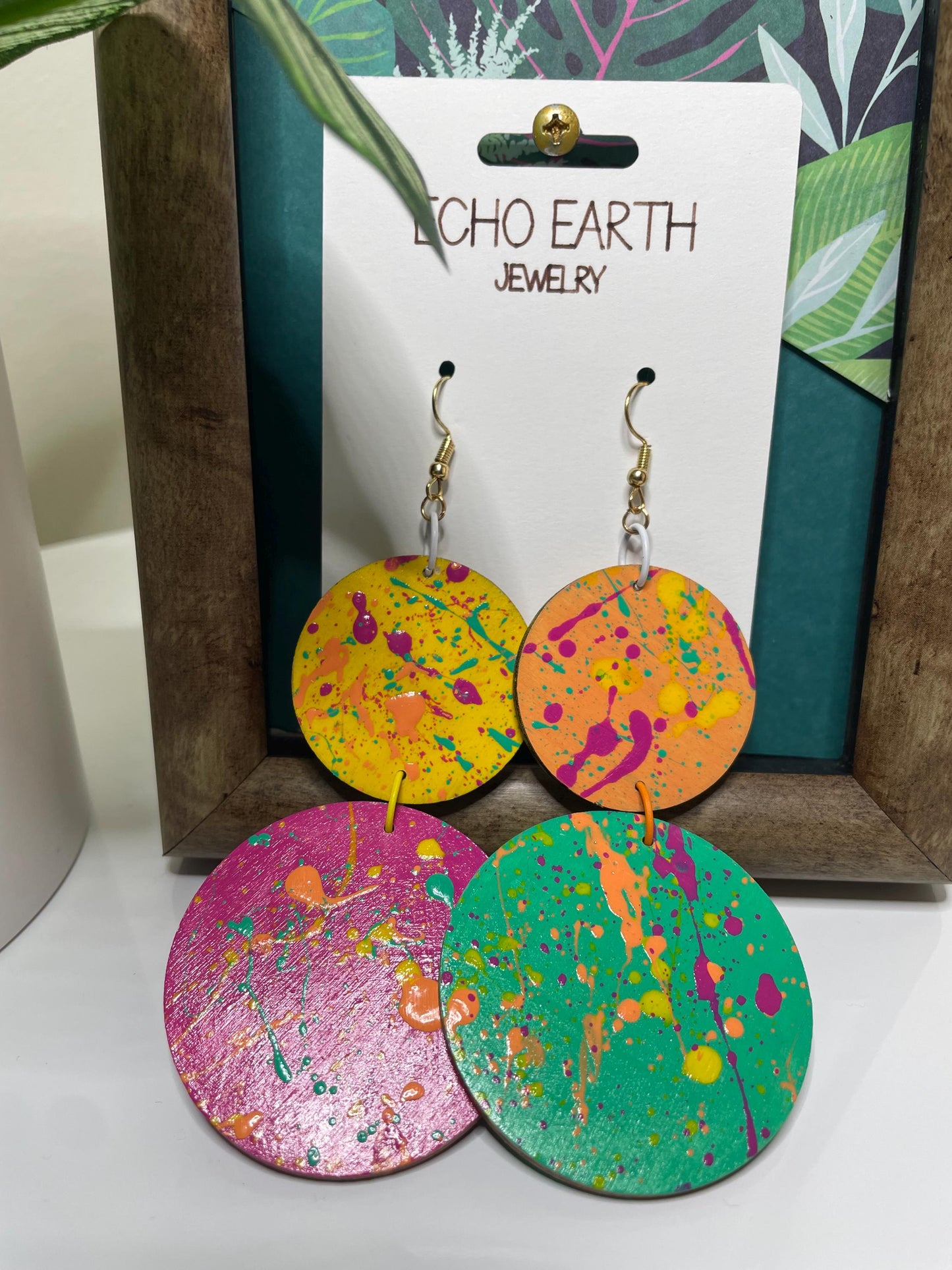 Colorful splatter-patterned earrings on a 'Echo Earth Jewelry' card with a wooden frame and plant in the background.